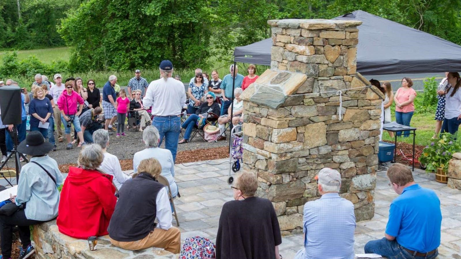 A group of people sit on a low stone wall behind a sandstone chimney monument. 