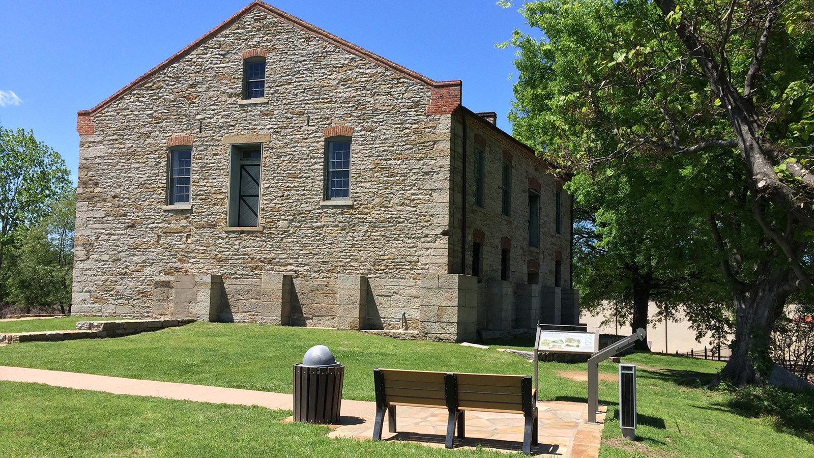 A bench and two wayside pannels in front an gray stone two story building on a sunny day.