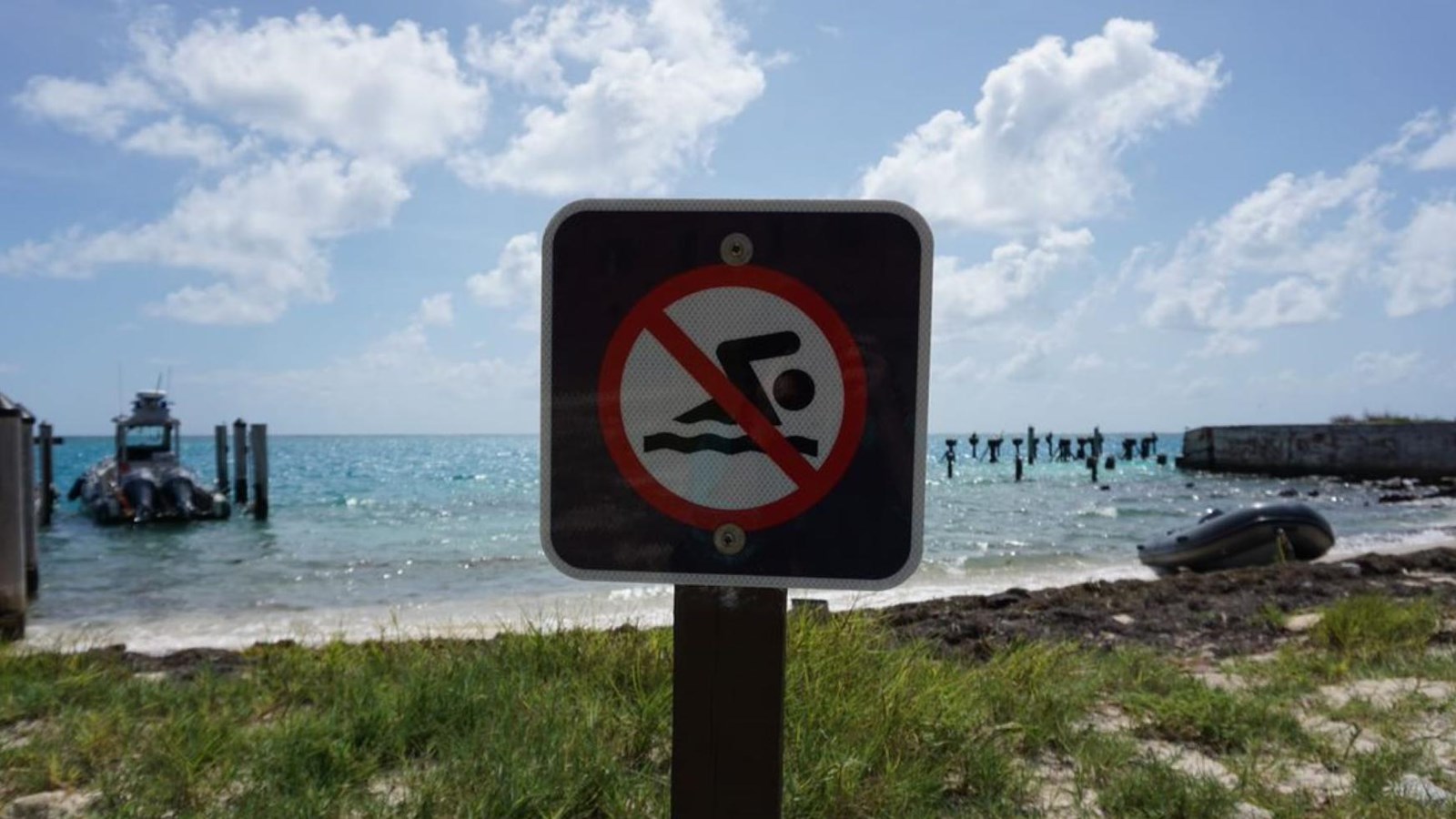 A park service sign indicating no swimming, a dinghy on a beach with blue water is the background.