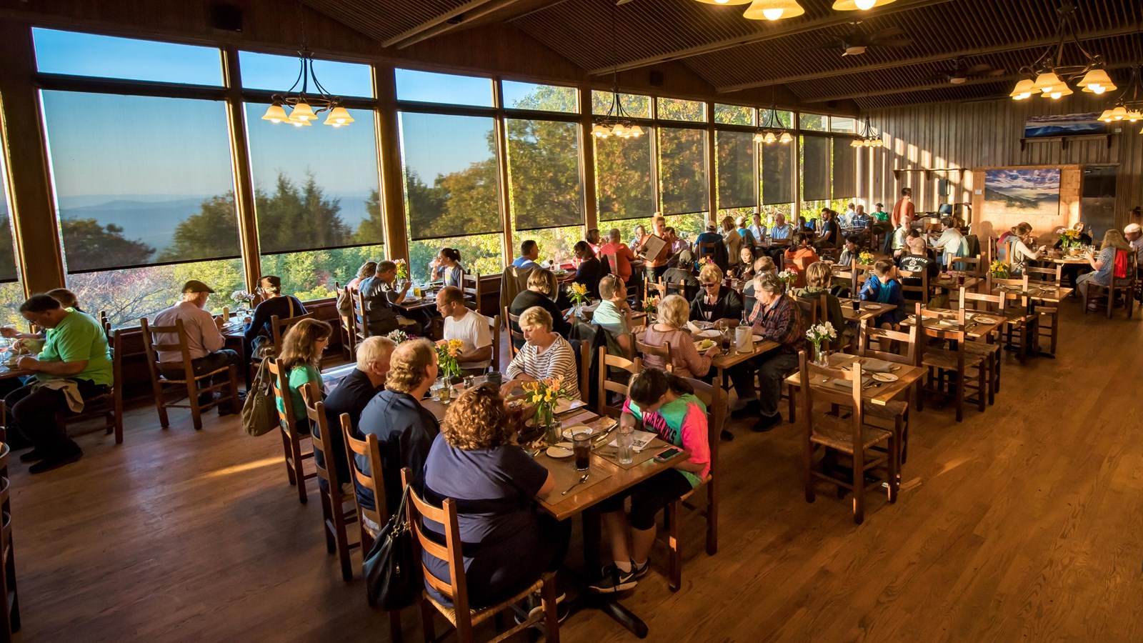 A group of diners sit at several tables in a dining room with wooden floors and large windows.