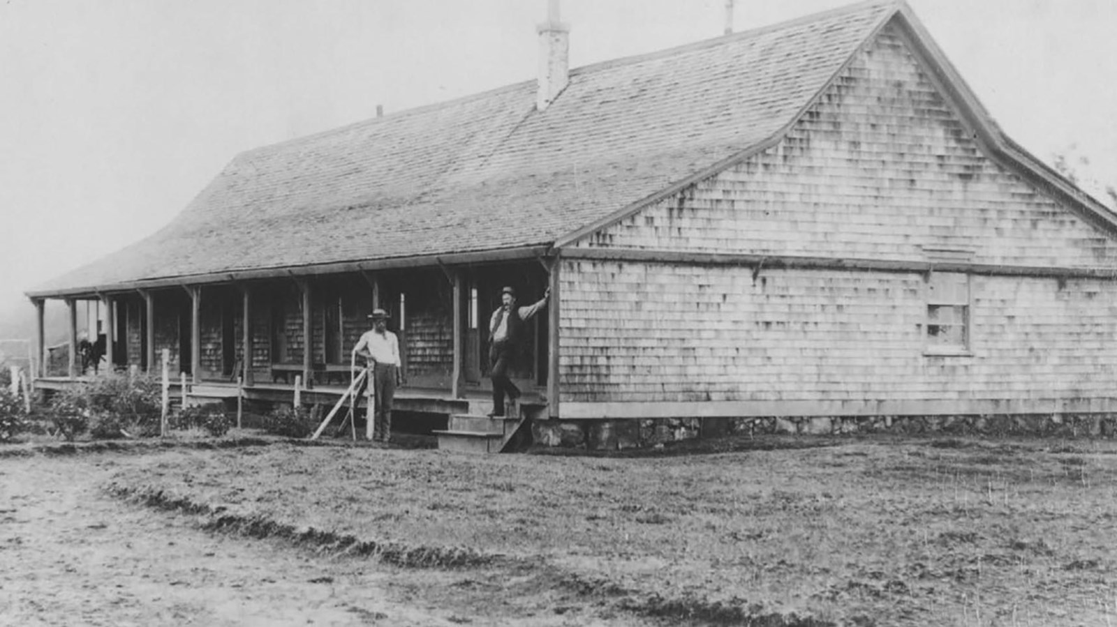 A black and white photo of a wooden building with two men standing on the porch