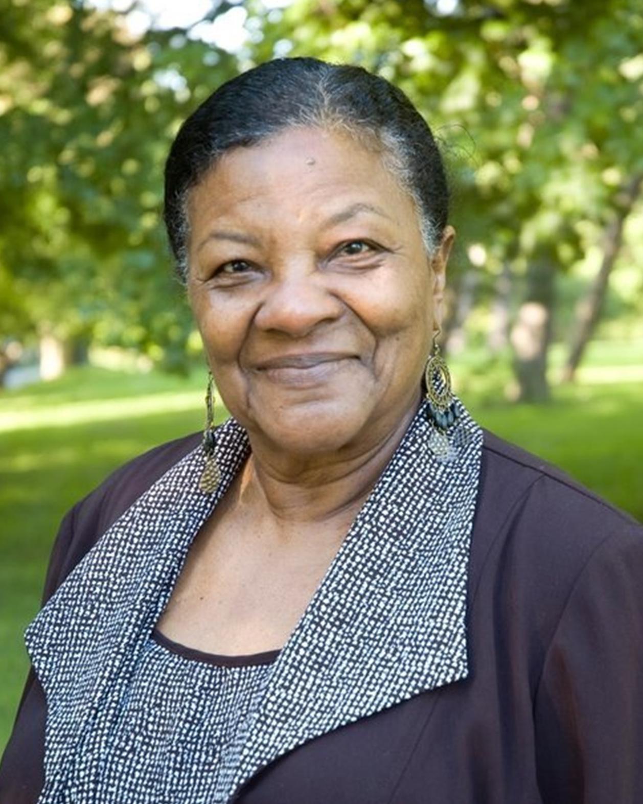 An older Black woman poses for a photo in a springtime park.