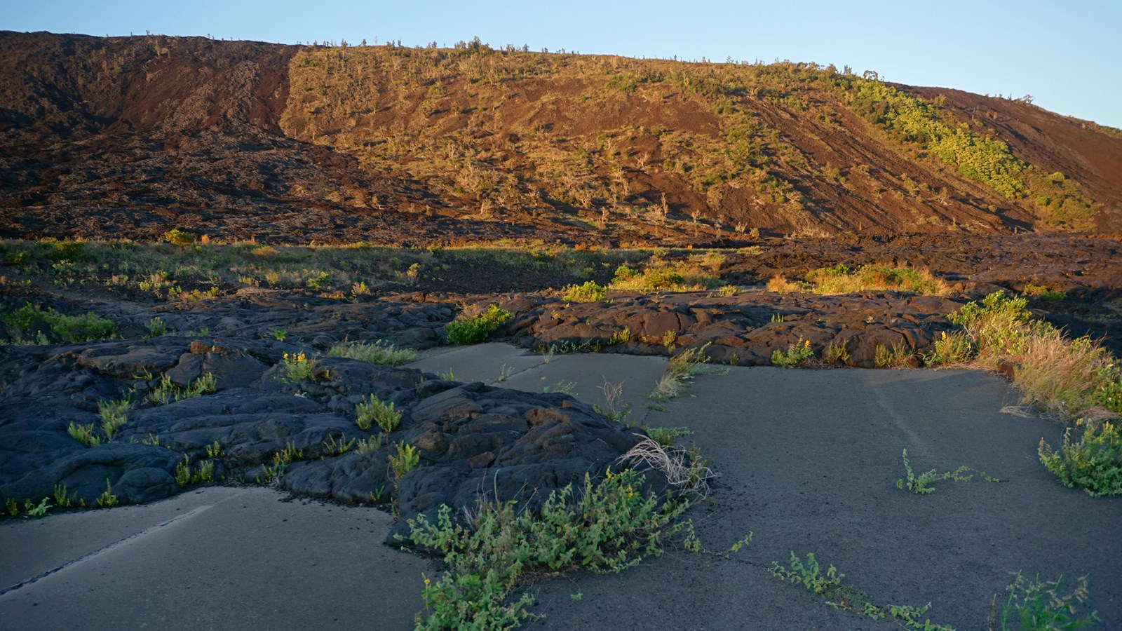 A road partially covered by black solidified lava at sunset with a steep hill in the background