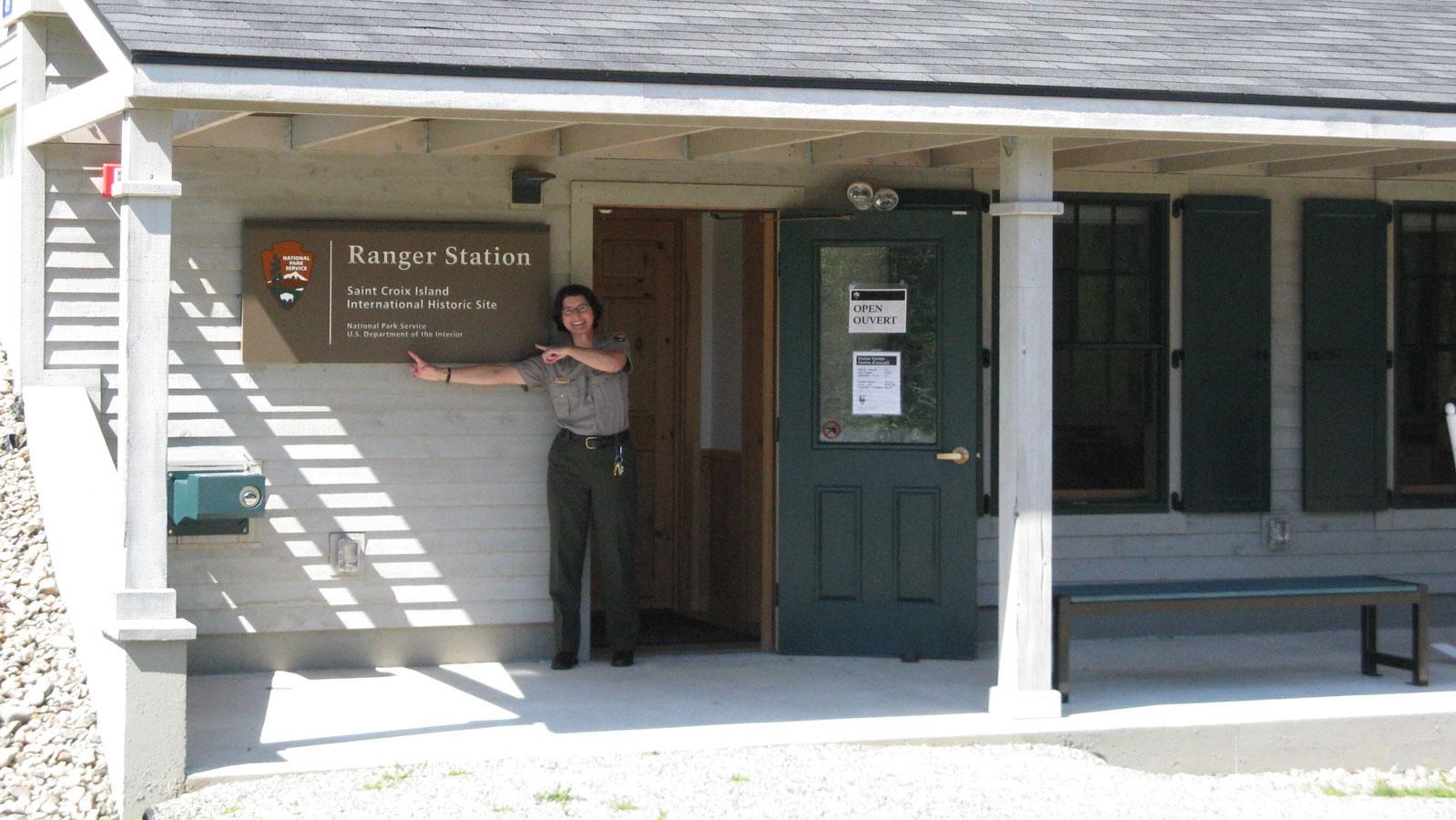 a ranger in front of \'ranger station\' sign waves hello in front of grey building with green shutters