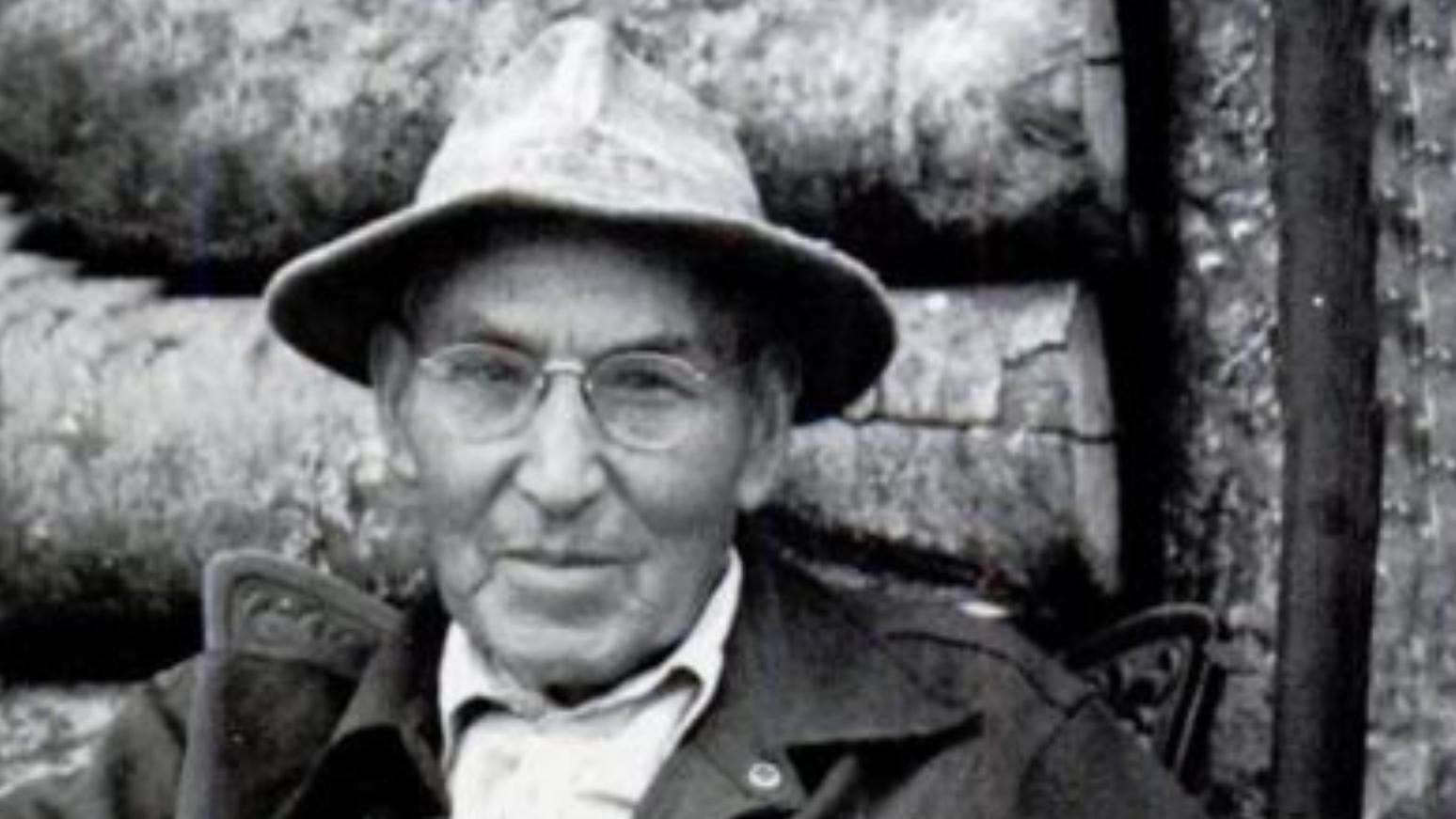 Black and white photograph of a seated elderly man wearing a fishing hat and holding a walking stick