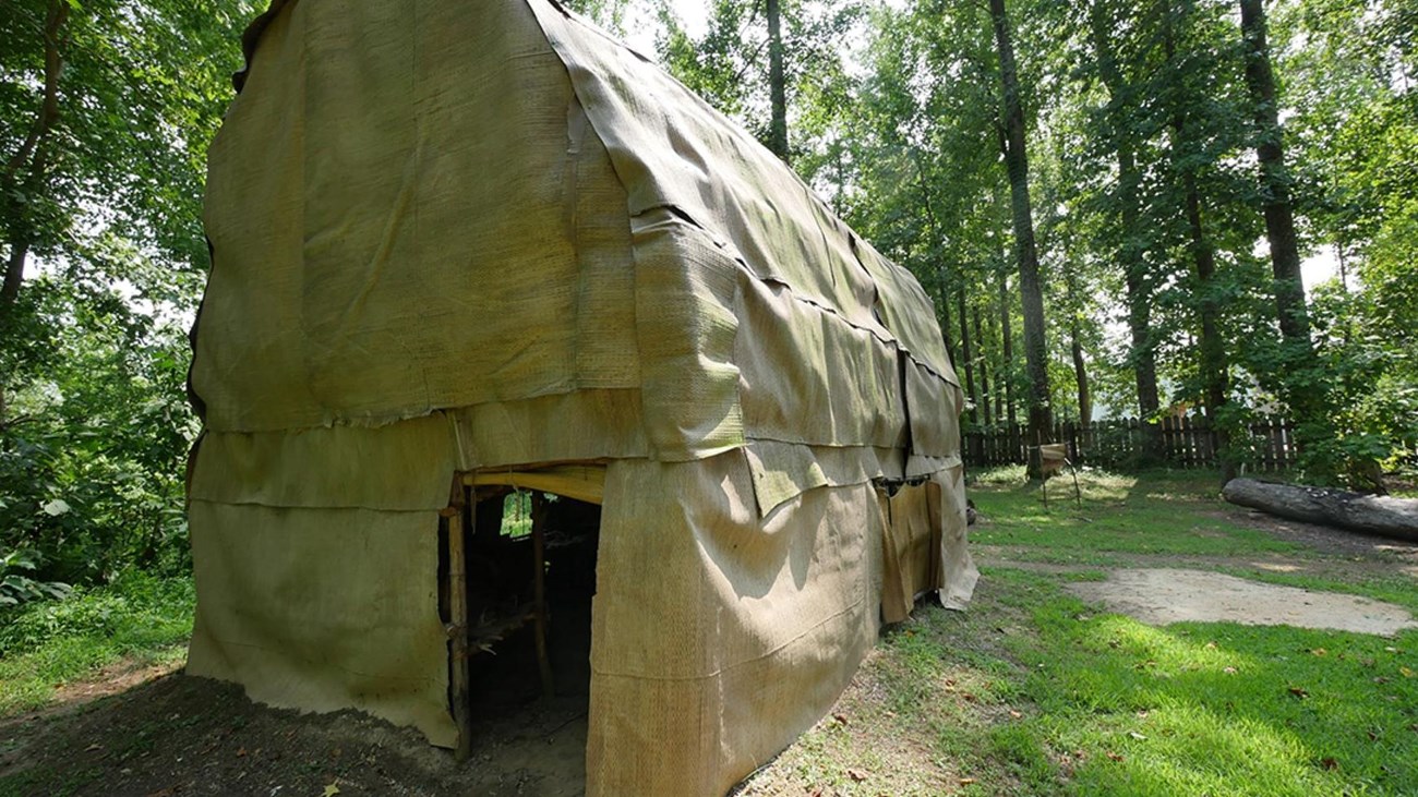 A Native American longhouse sitting in a grassy area under trees.