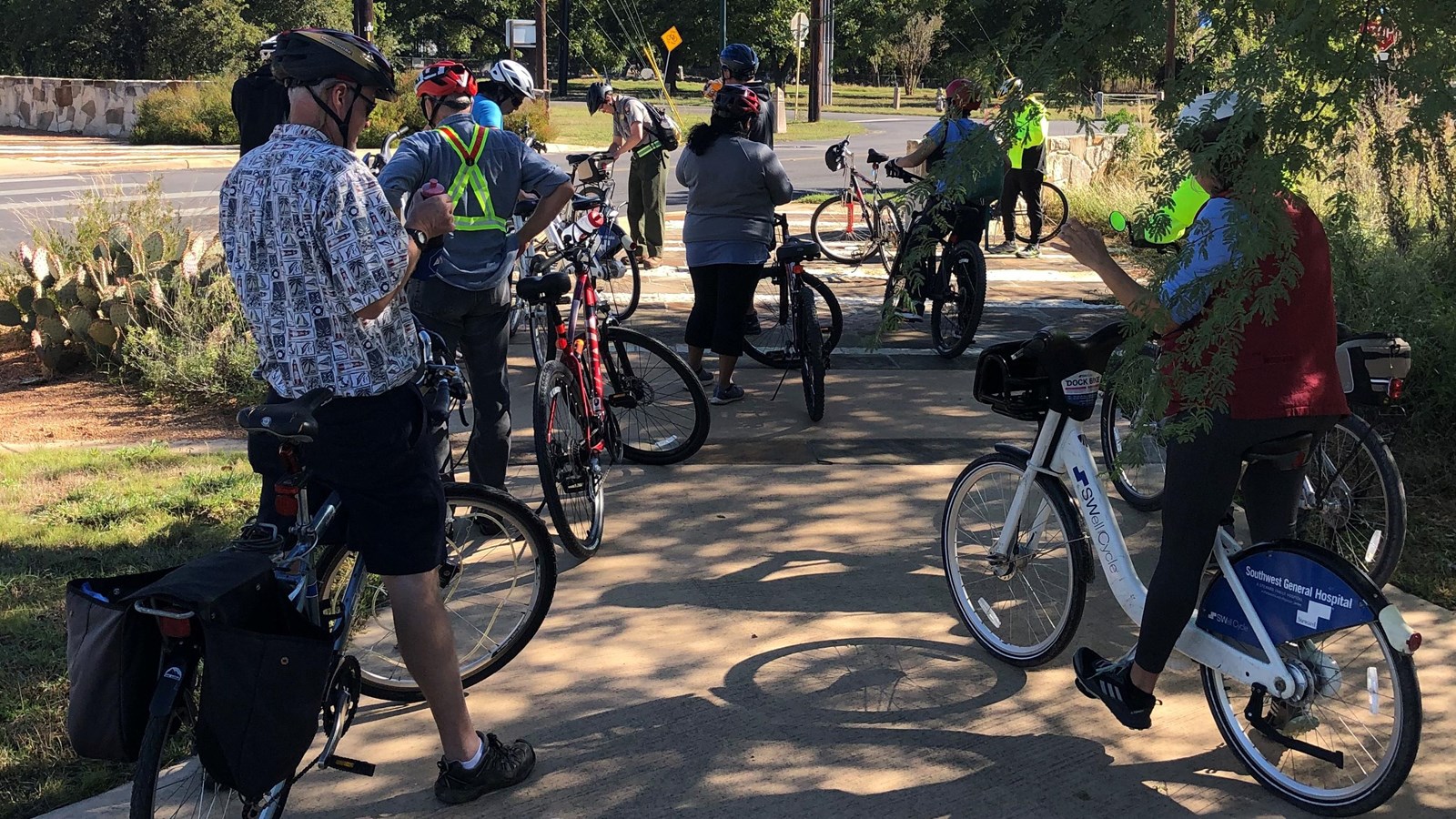 Group of bicyclists pause under the shade of a tree on the paved trail.