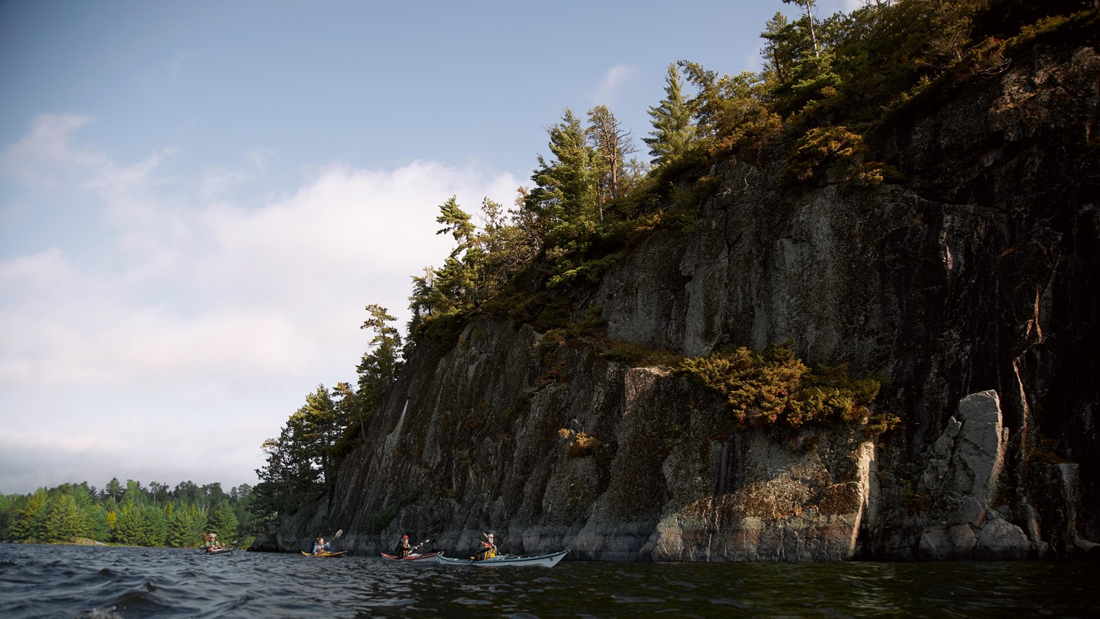 People paddle in kayaks by cliffs that raise 125 feet out of the water. Pine trees grow on cliff top