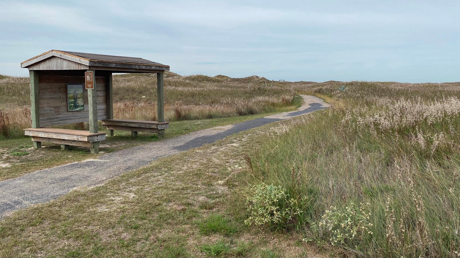 A paved trail leads into a wide-open prairie. A small shelter with benches is next to the trail.