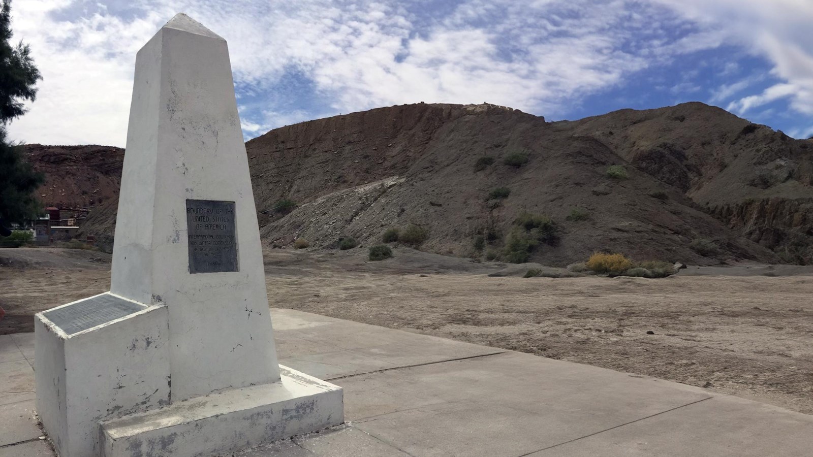 A white obelisk with a metal plaque, surrounded by mountainous terrain and desert vegitation.