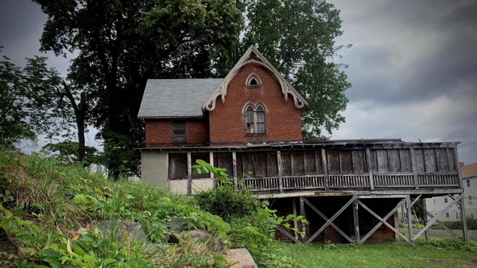A red, multistory brick building with an enclosed porch nestled underneath a tree. 