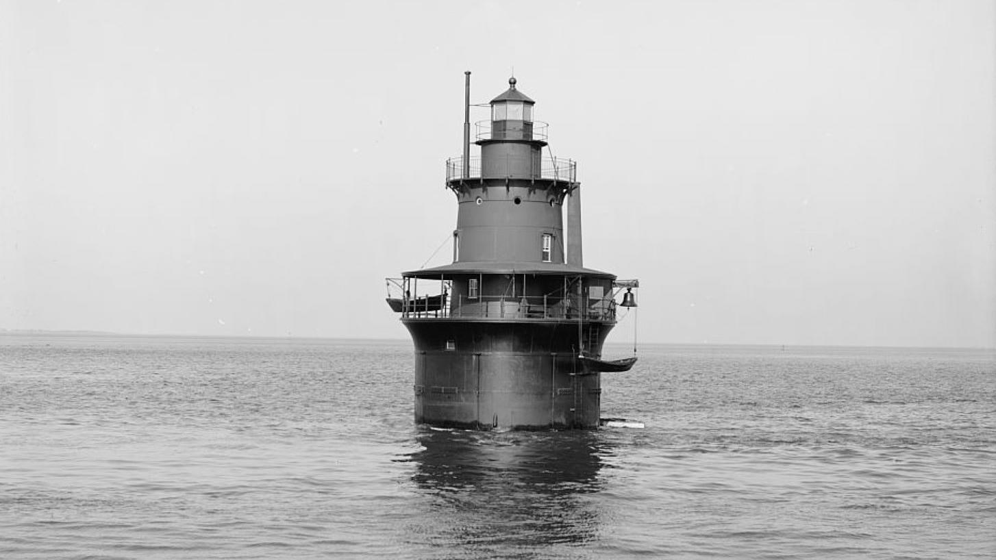 Round, short, metal buoy lighthouse next to a small cropping of rocks in water. 