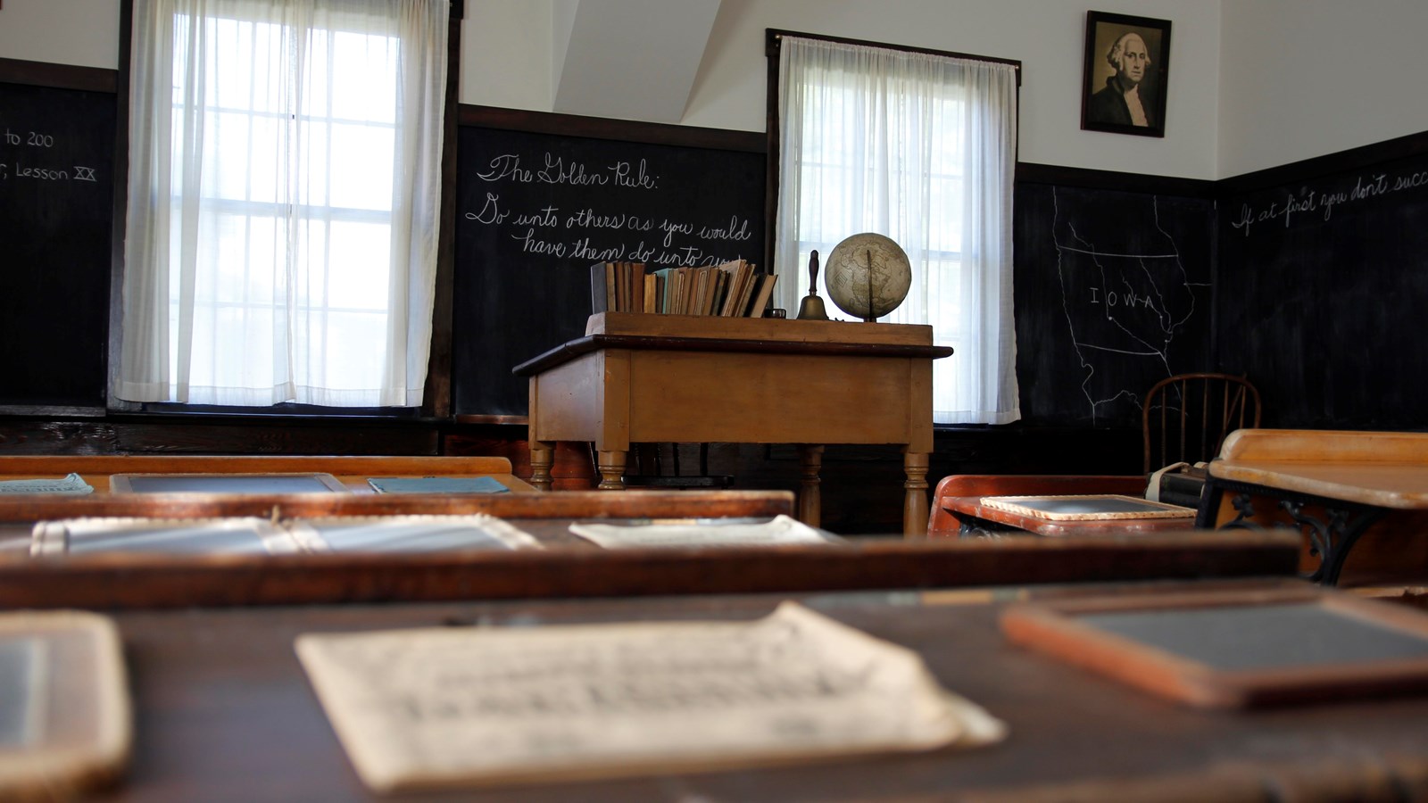 An 1870s classroom with an elevated teacher\'s desk and chalkboards as seen from a pupil\'s desk.