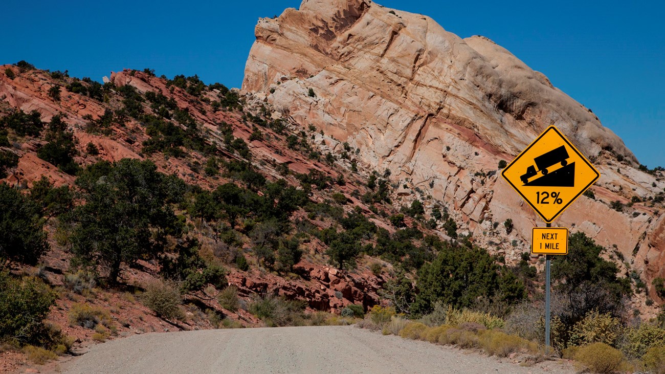 Dirt road that curves around a large rock. Road sign indicating steep incline ahead.