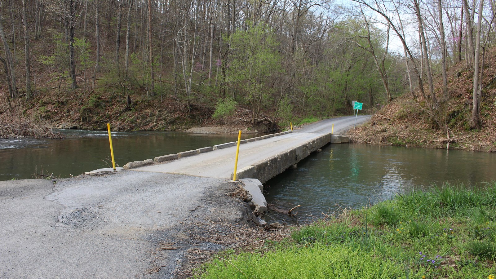 A country road crosses a woodland creek on a low, one-lane concrete bridge.