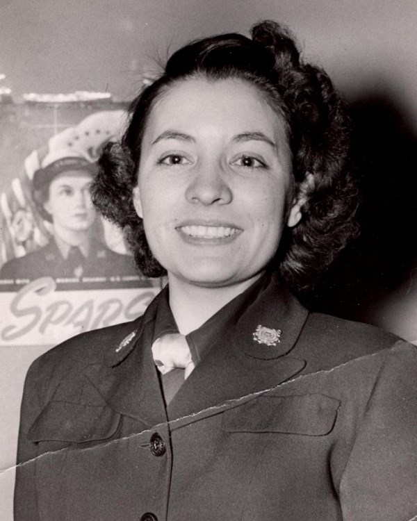 Young woman smiling at the camera in dark military jacket. A SPARS propaganda poster is behind her. 