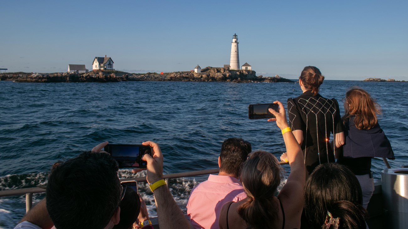 People on a boat taking pictures or looking at a white lighthouse on a rocky island.