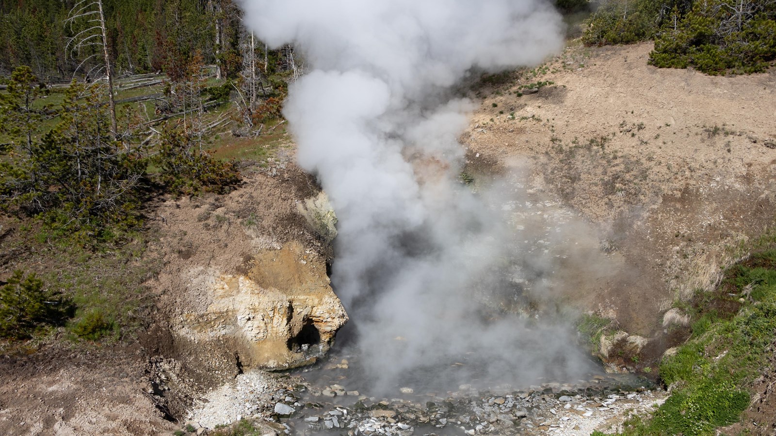Steam rises out of a cave from the waters of a hot spring located inside.