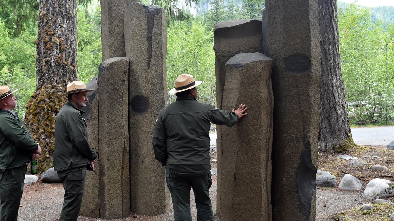 Three park rangers stand next to a stone memorial made of seven basalt columns