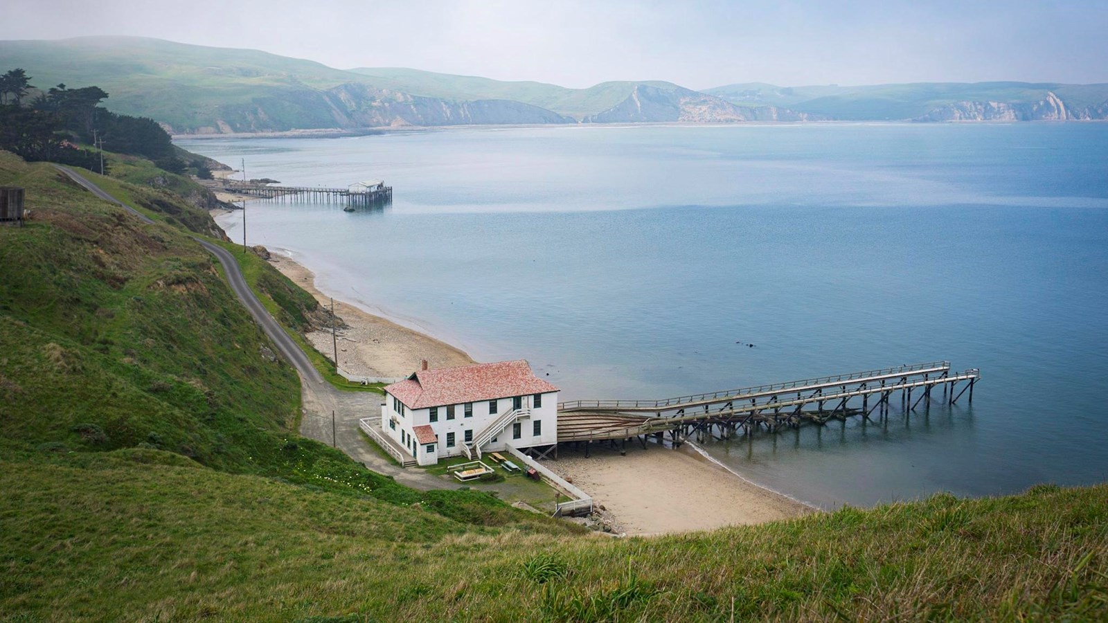 The historic boathouse sits in the shelter of Drakes Bay. A railway leads out to the water.