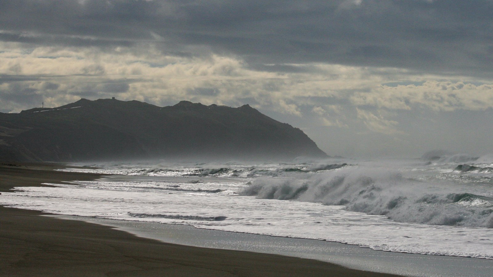 Large waves crash onto a sandy beach on a mostly cloudy day. Rocky headlands rise in the background.
