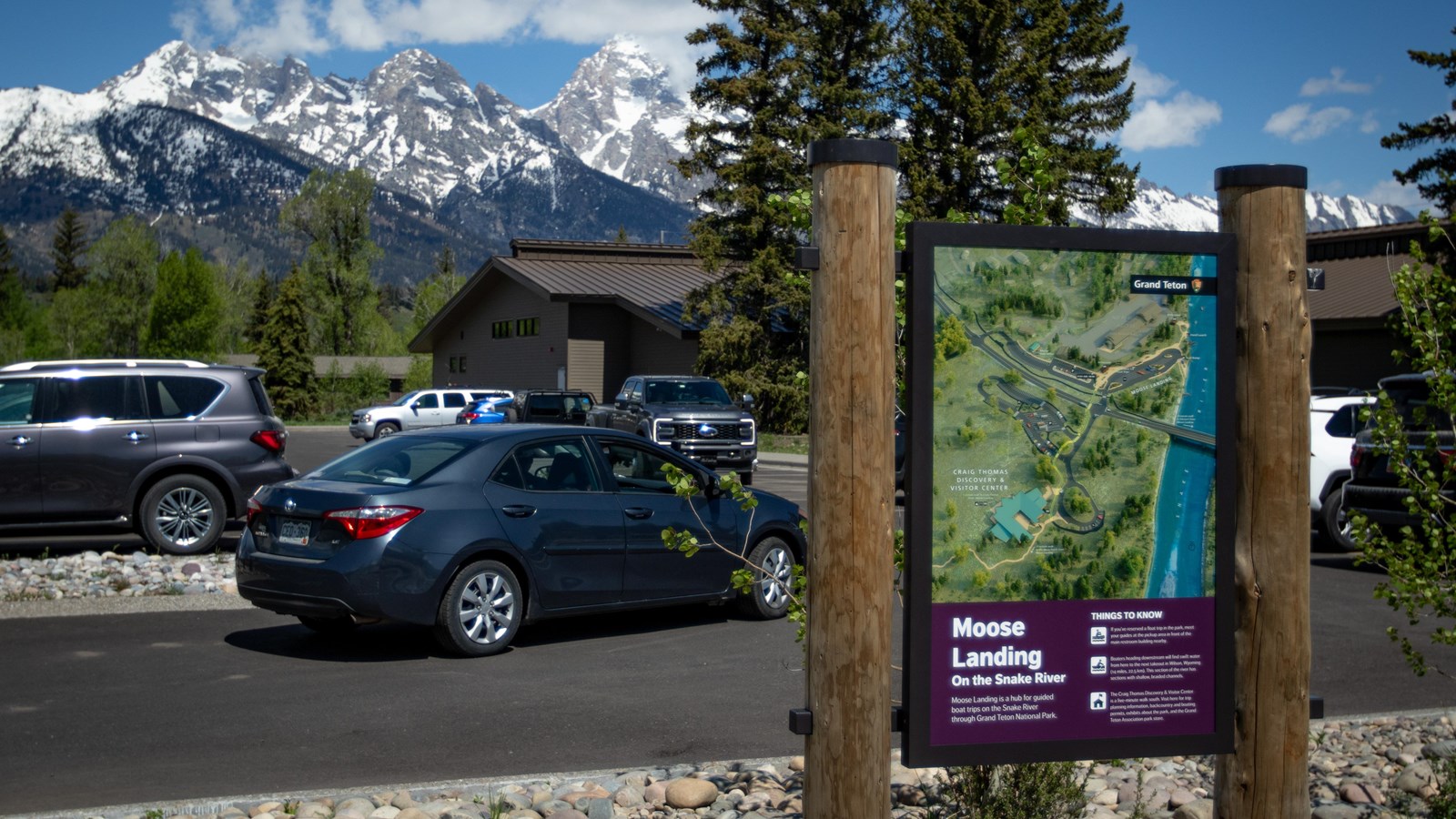 An informational sign in front of a parking lot with a car driving by and mountains in the back. 