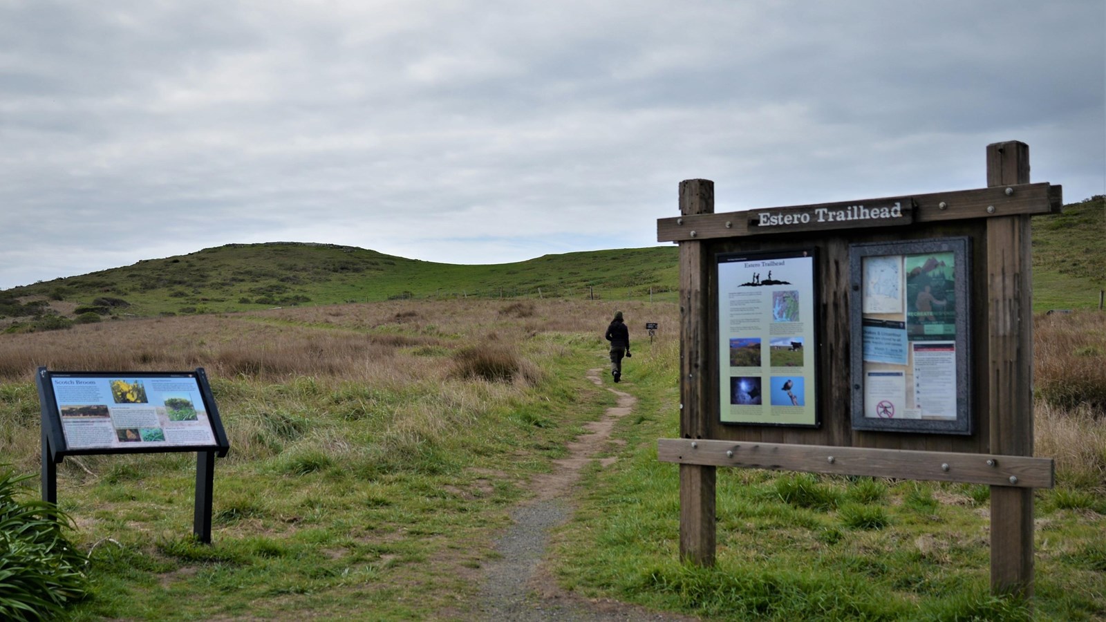 A hiker heads up a dirt trail between two signs under cloudy skies over green grassy hills.