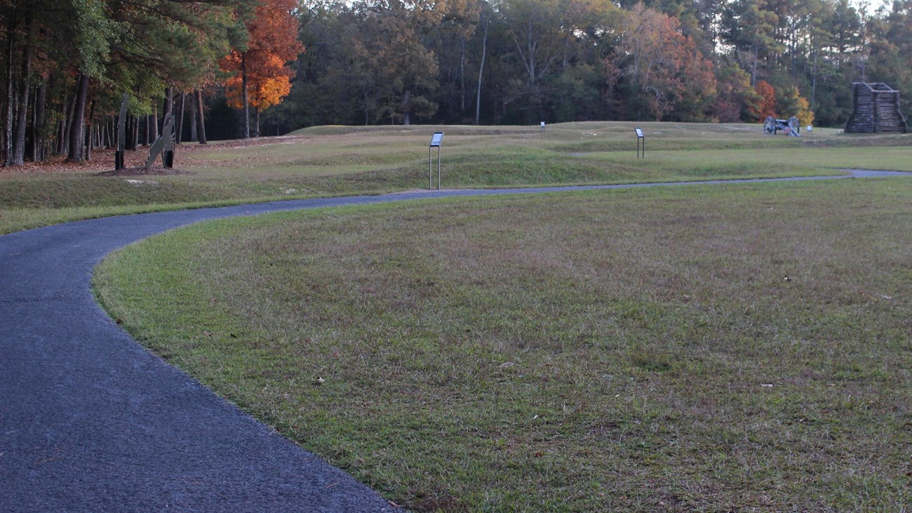 A paved trail winds through a field, past signs and toward a cannon and wooden tower. 