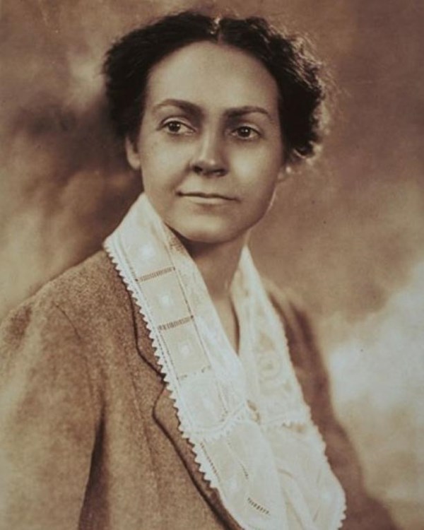 A younger woman with her dark hair up and parted down the middle. Wearing a suit with a lace scarf.