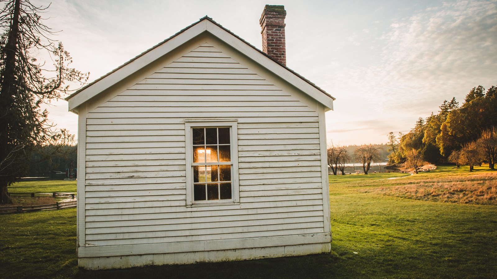 Color photograph of a white building made of wooden boards. The sunset is shining through it