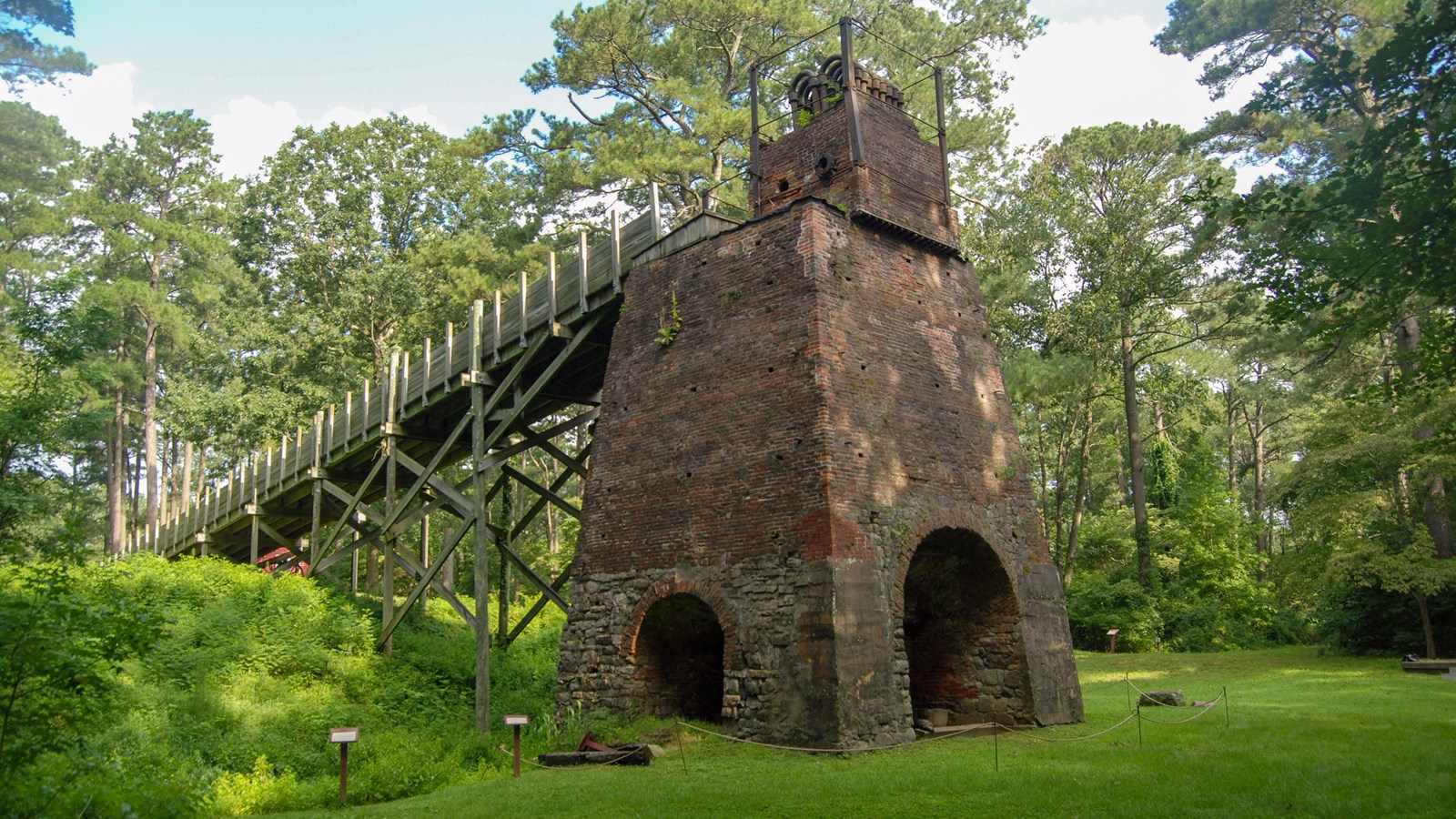 Old brick and stone furnace with a wooden walkway in a wooded area.