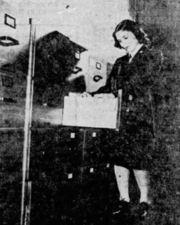 Grainy photo of woman in a dark skirt suit going through files at a wall of file cabinets. 