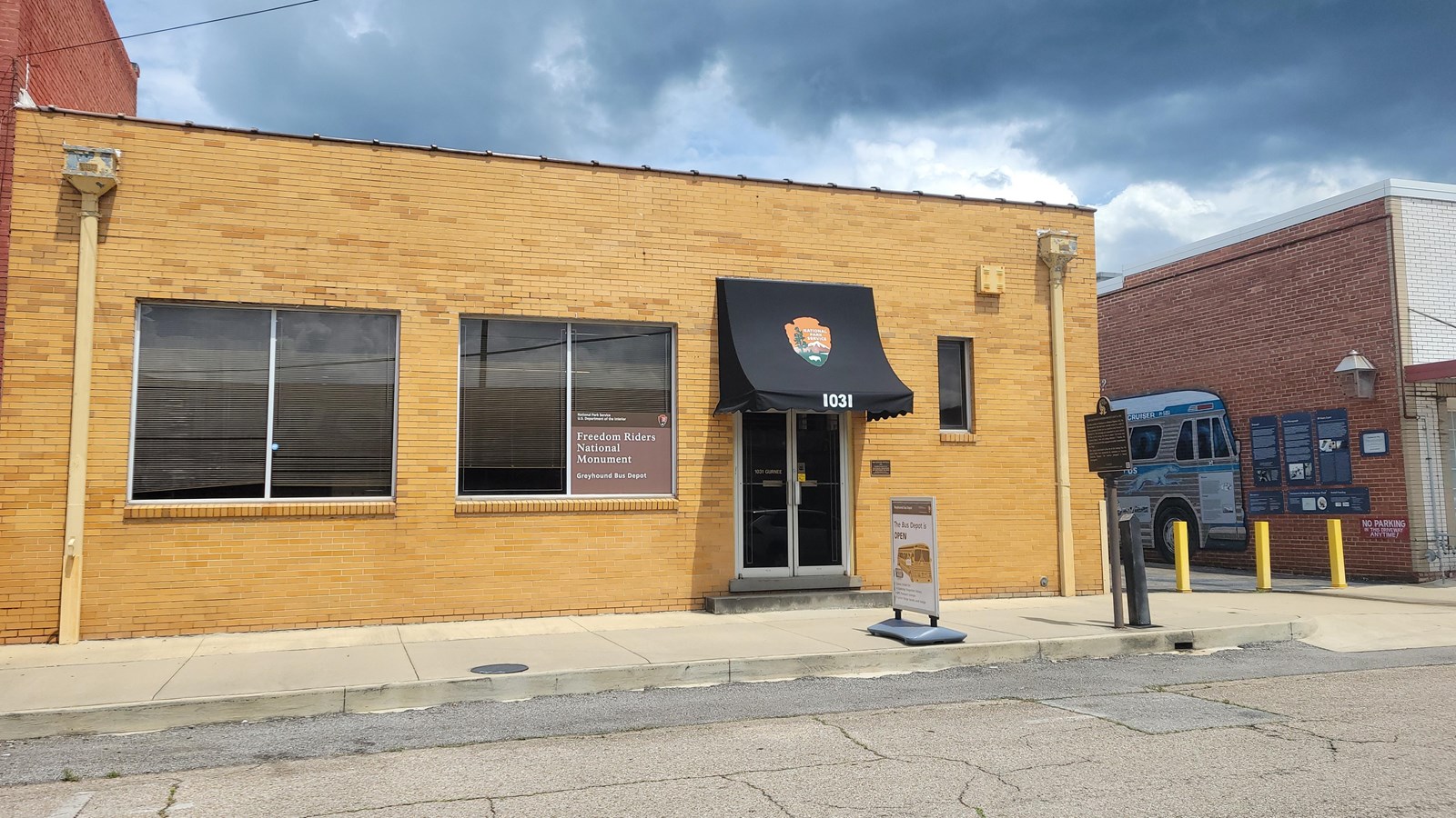 A tall one story brick building with large windows. An alley next to the building has a bus mural.