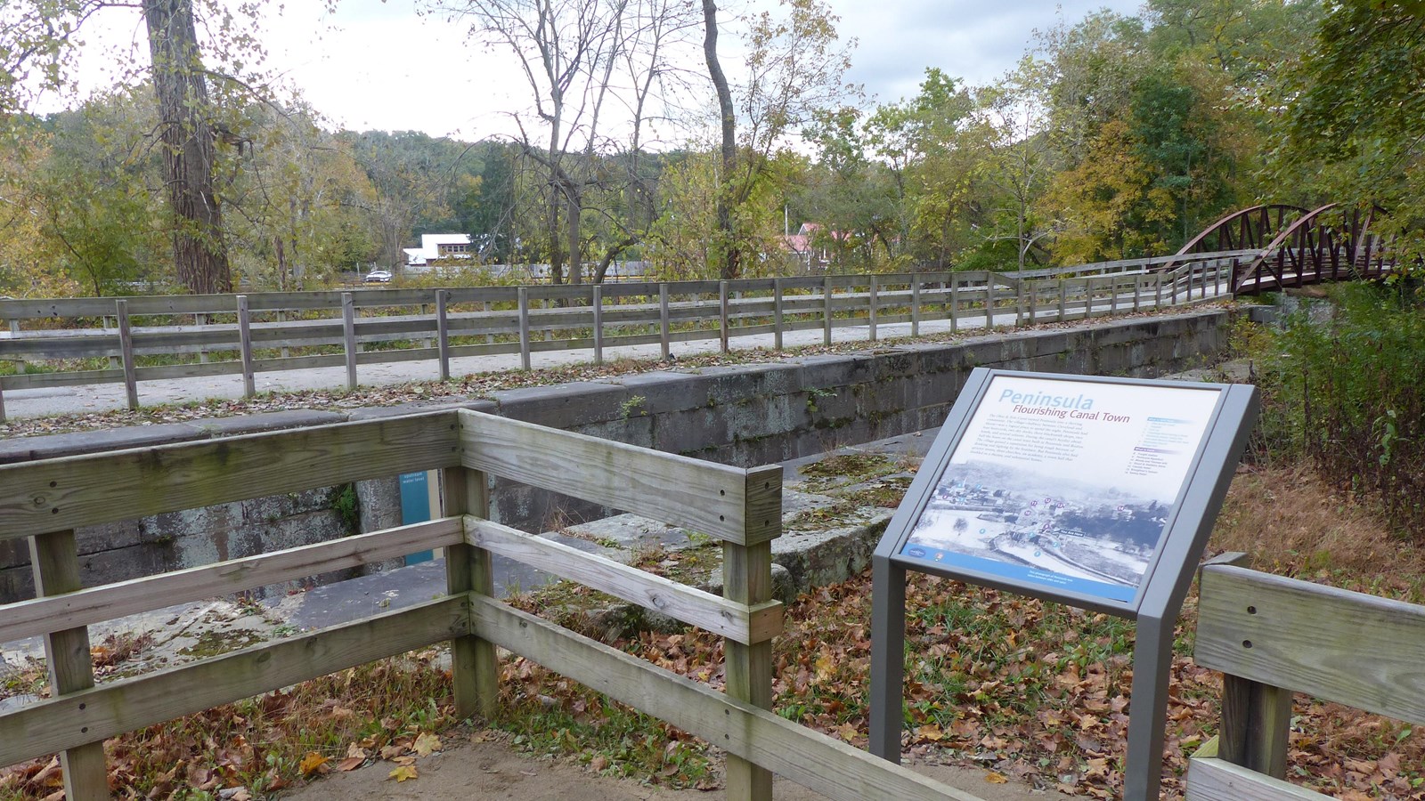 An exhibit panel along a wooden fence overlooking two parallel stone walls next to a trail.