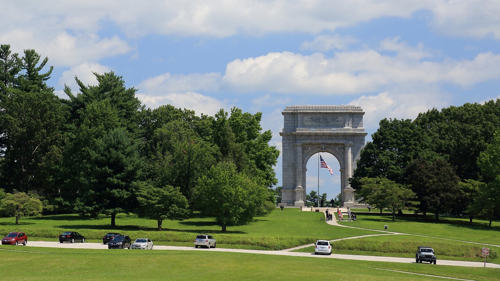 photograph of large stone arch monument with american flag, grassy lawn and parking and pathways