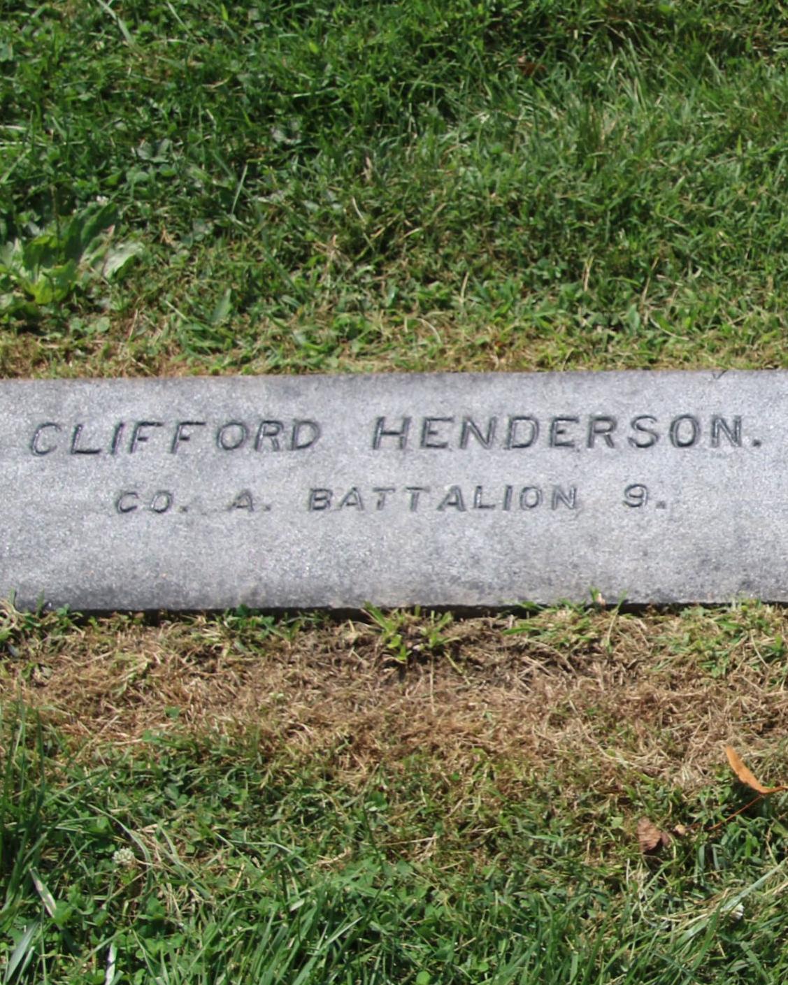 A dark colored gravestone engraved with the name Clifford Henderson lays flat among grasses. 