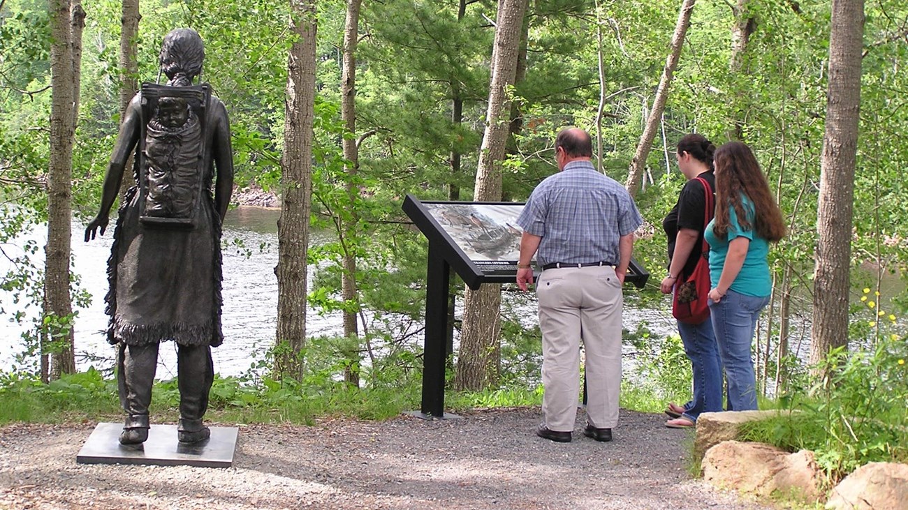 Three people stand in front of a wayside next to a bronze statue