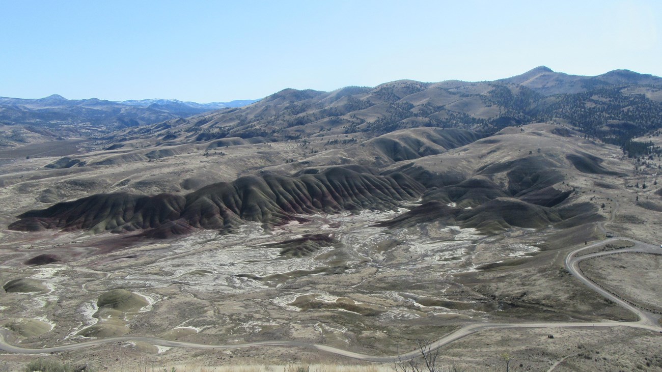 View of red and green striped hills from trail above, with green sagebrush and windy dirt road below