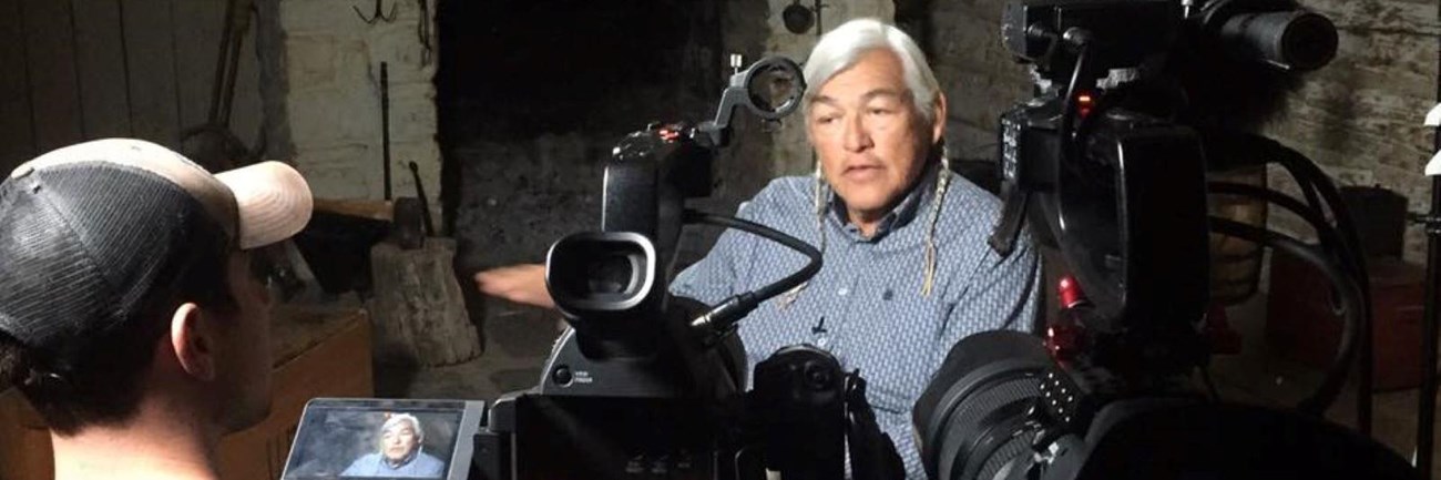One man speaks facing a camera, second man watched playback on TV monitor