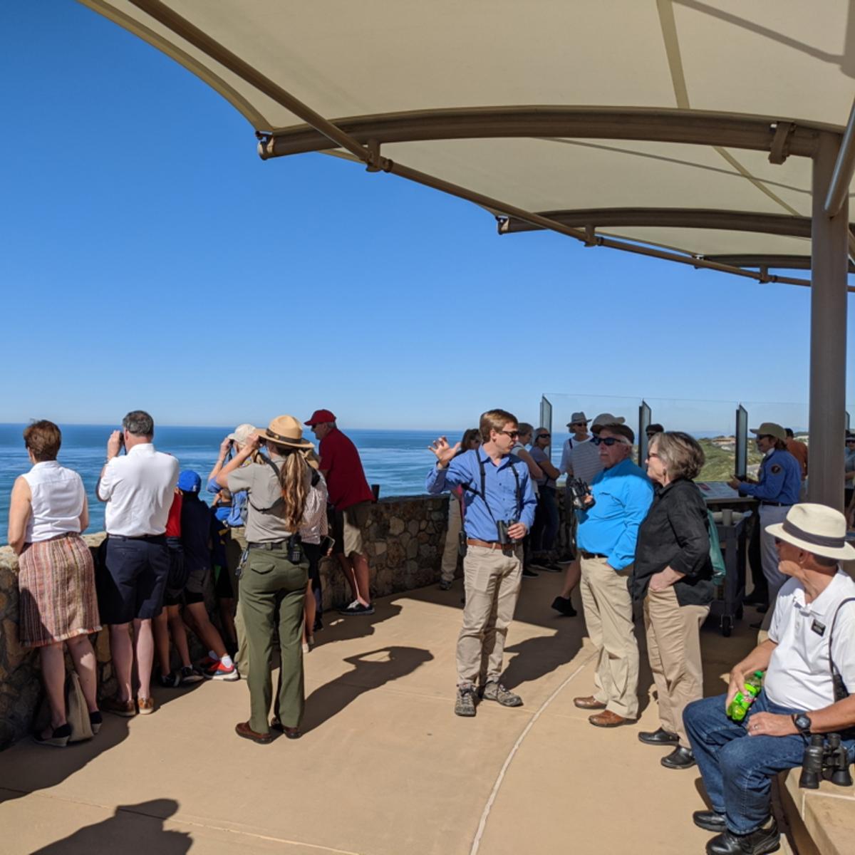 A crowd of people gather under a white, cloth covering. They look over a fence towards blue water.