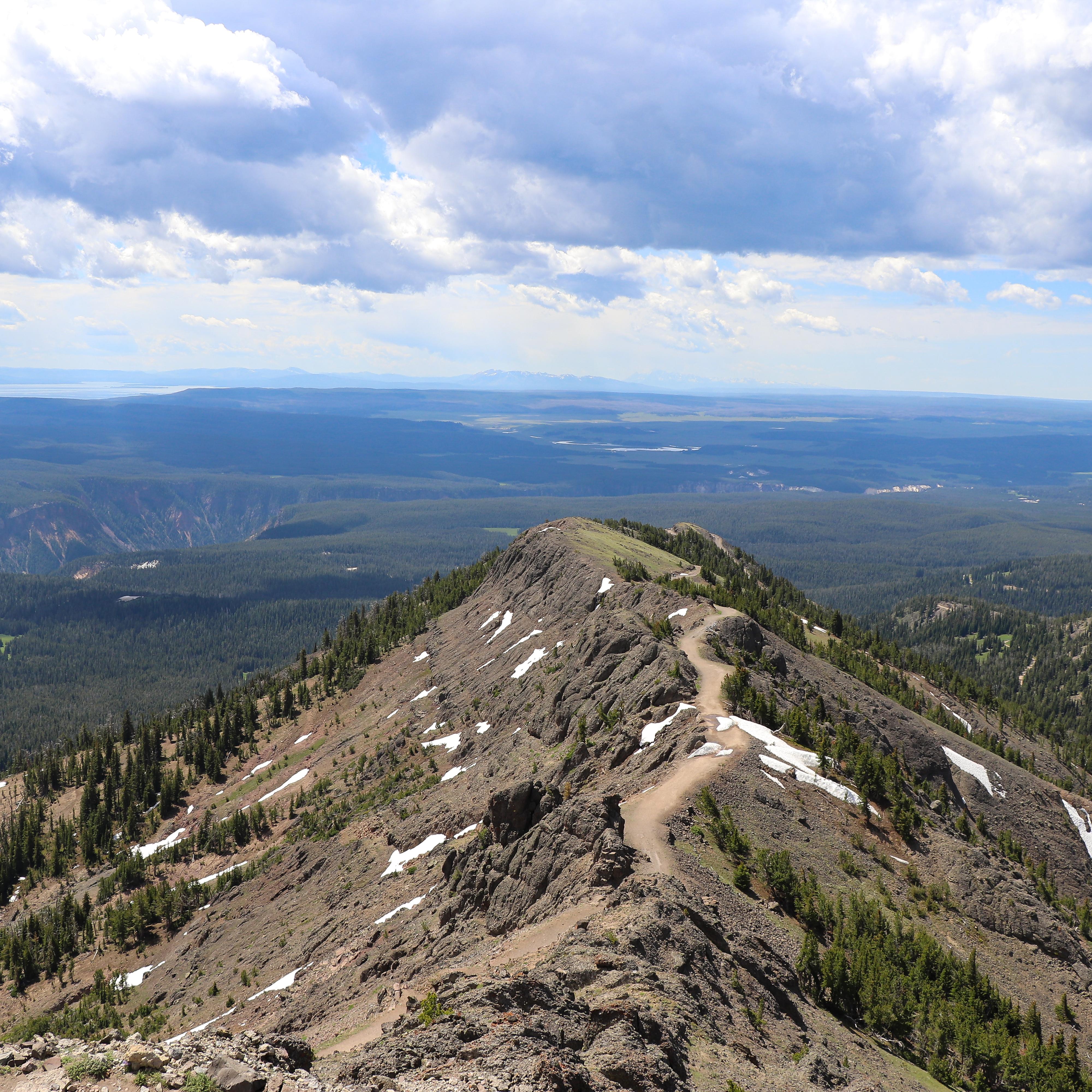 A trail crosses a ridgeline above forests and meadows.