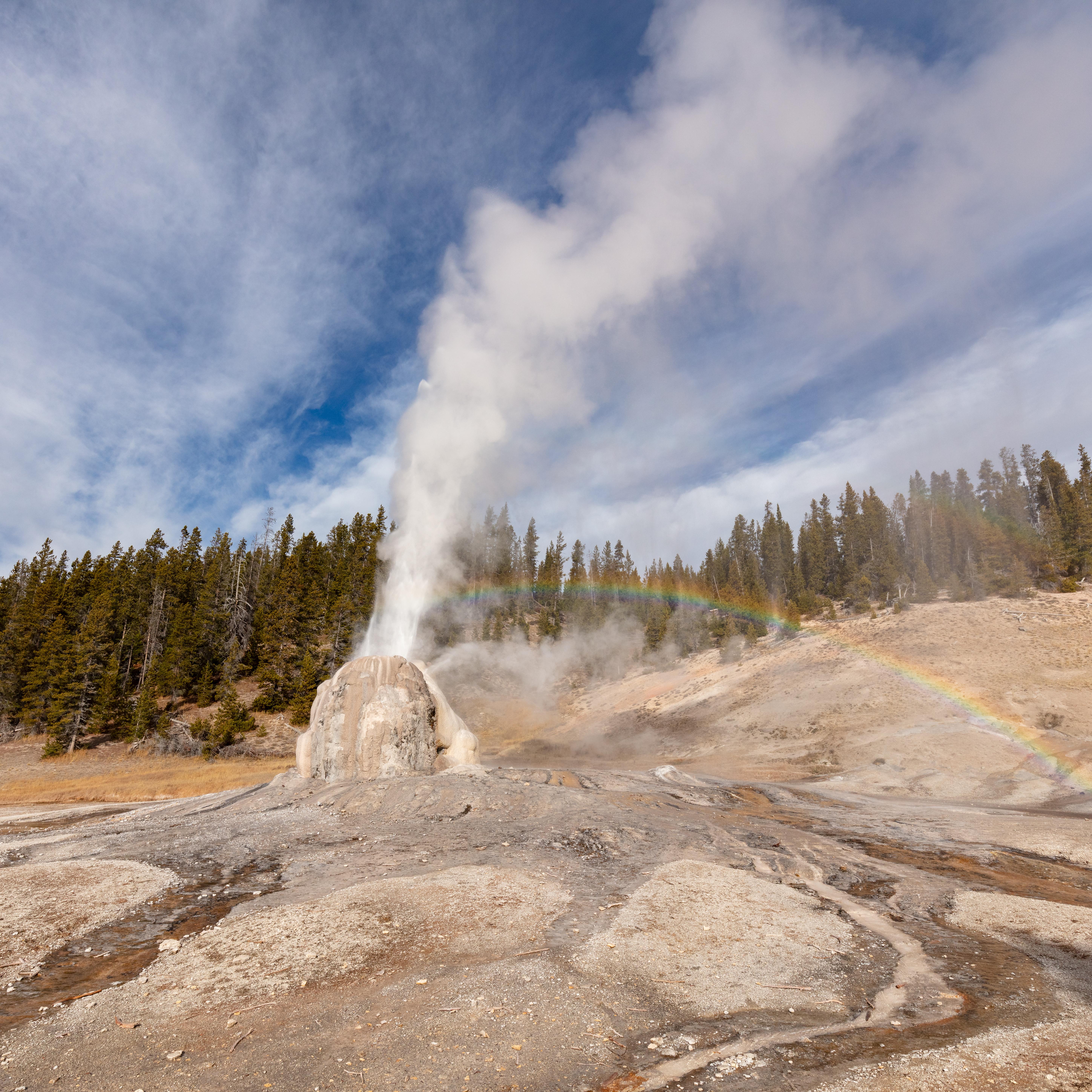 Steam and water erupt from the tan cone of Lone Star Geyser. A rainbow appears in the mist.