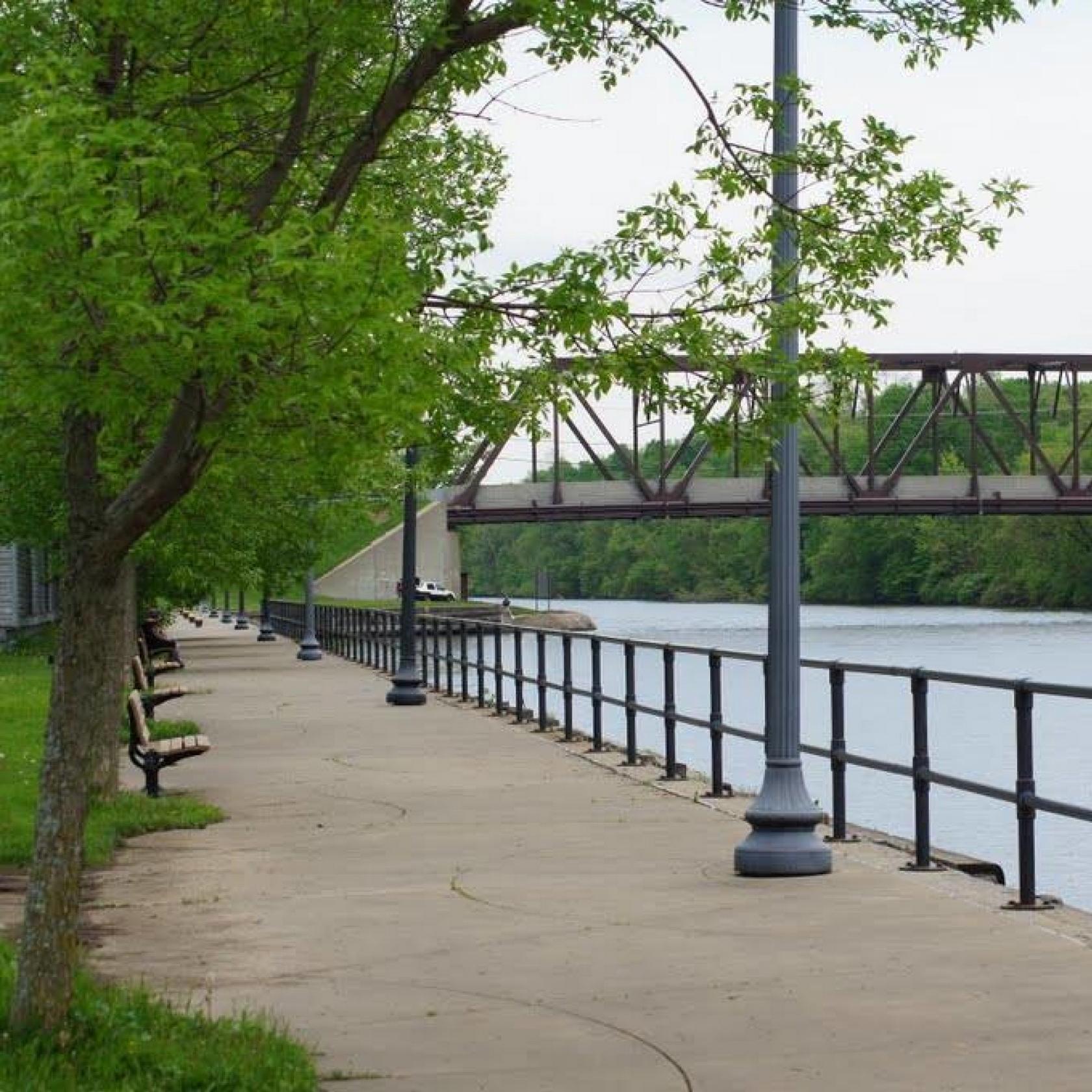 Along a sidewalk, you can see a metal railing and just beyond that, a river and a metal bridge. 