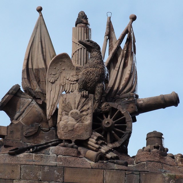 A red stone statue. It has carved banners and an eagle perched on the barrel of a carved cannon. 