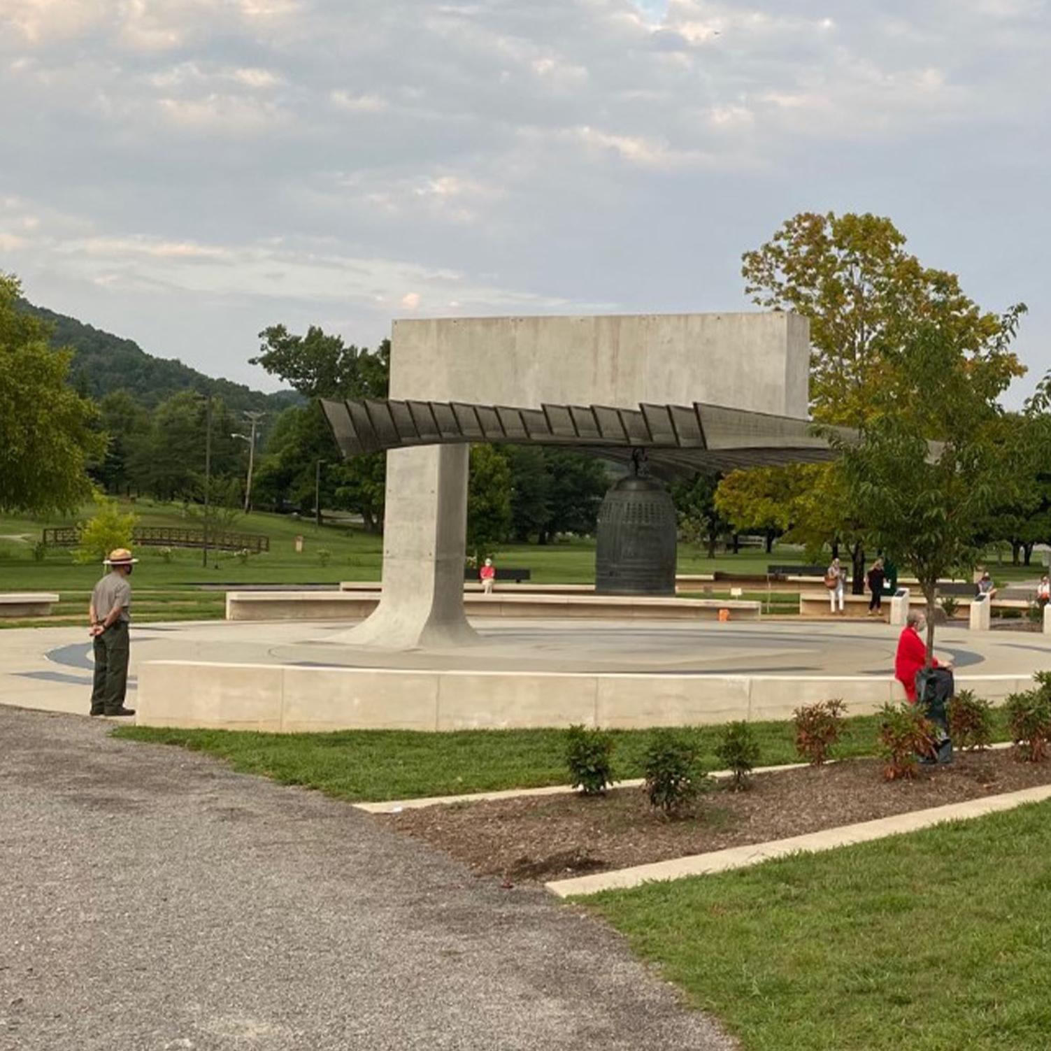 People gathered around a large bronze bell with grass, trees, and trails throughout