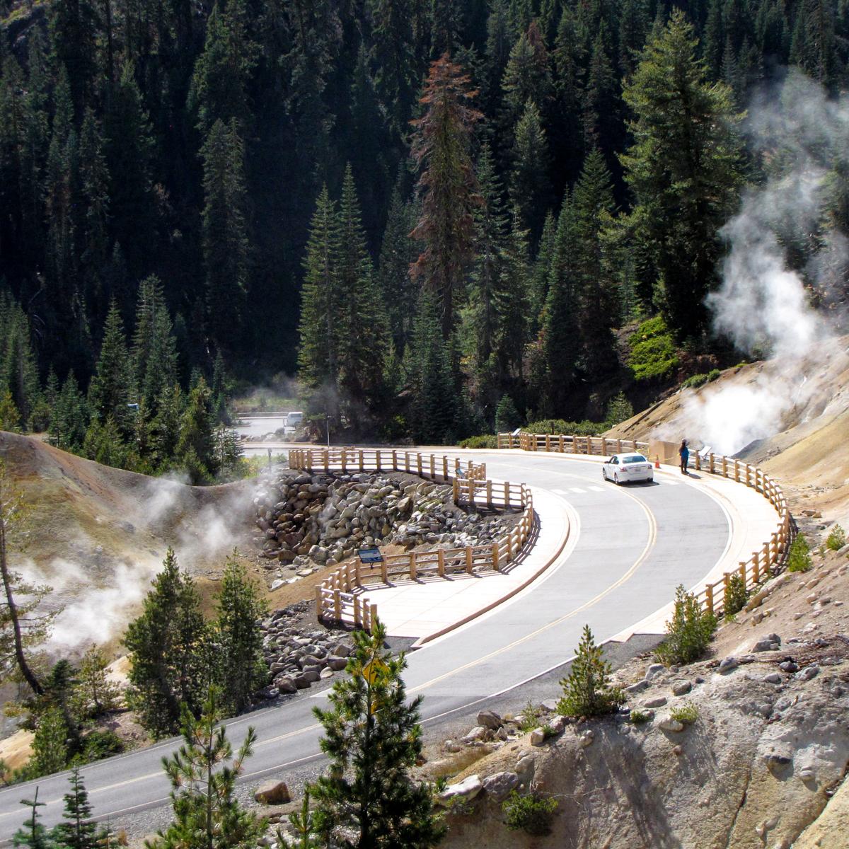 A road lined by wooden guardrails curves through a hydrothermal area of barren, steaming ground.