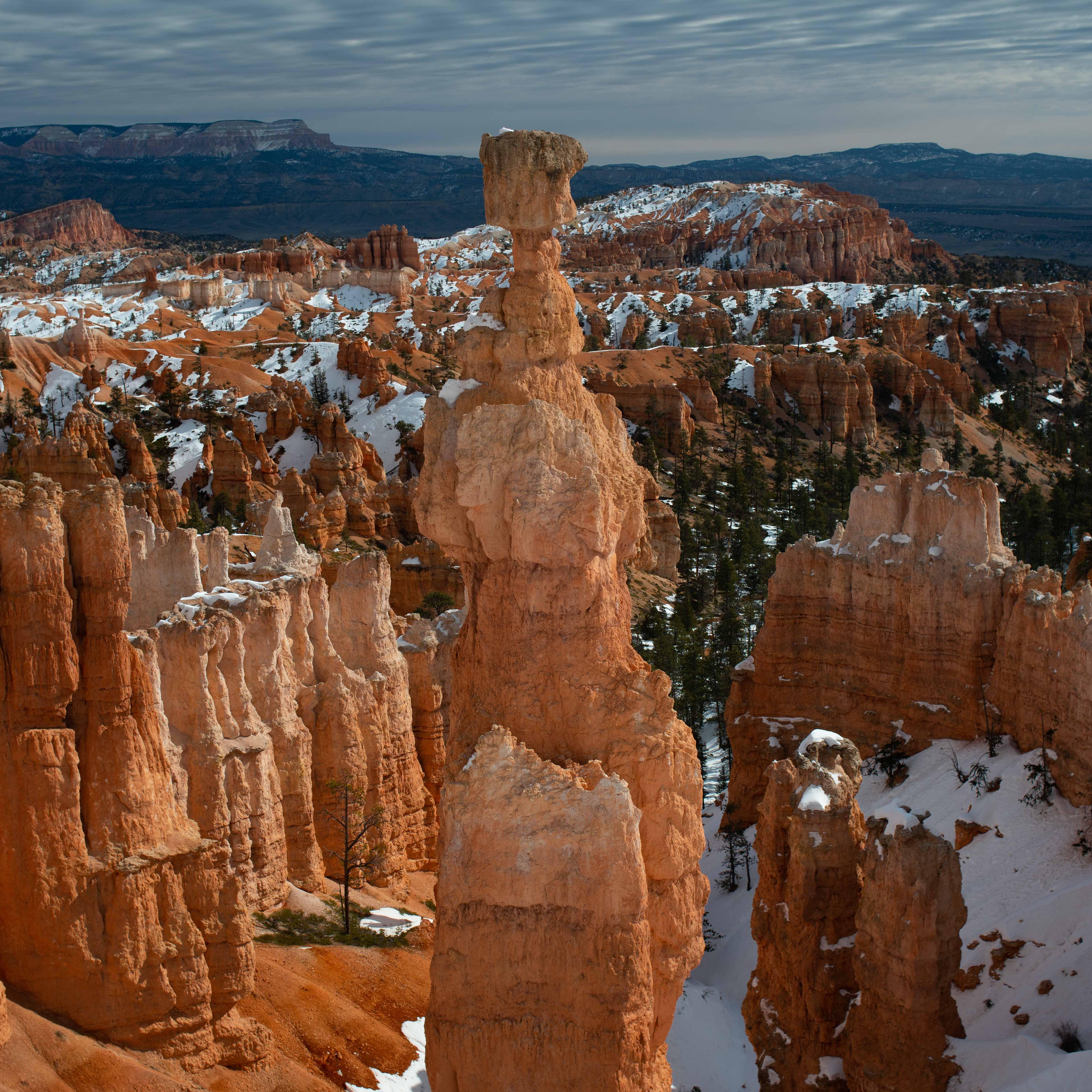 A tall spire of red rock stands against a background of stone, snow, and overcast skies