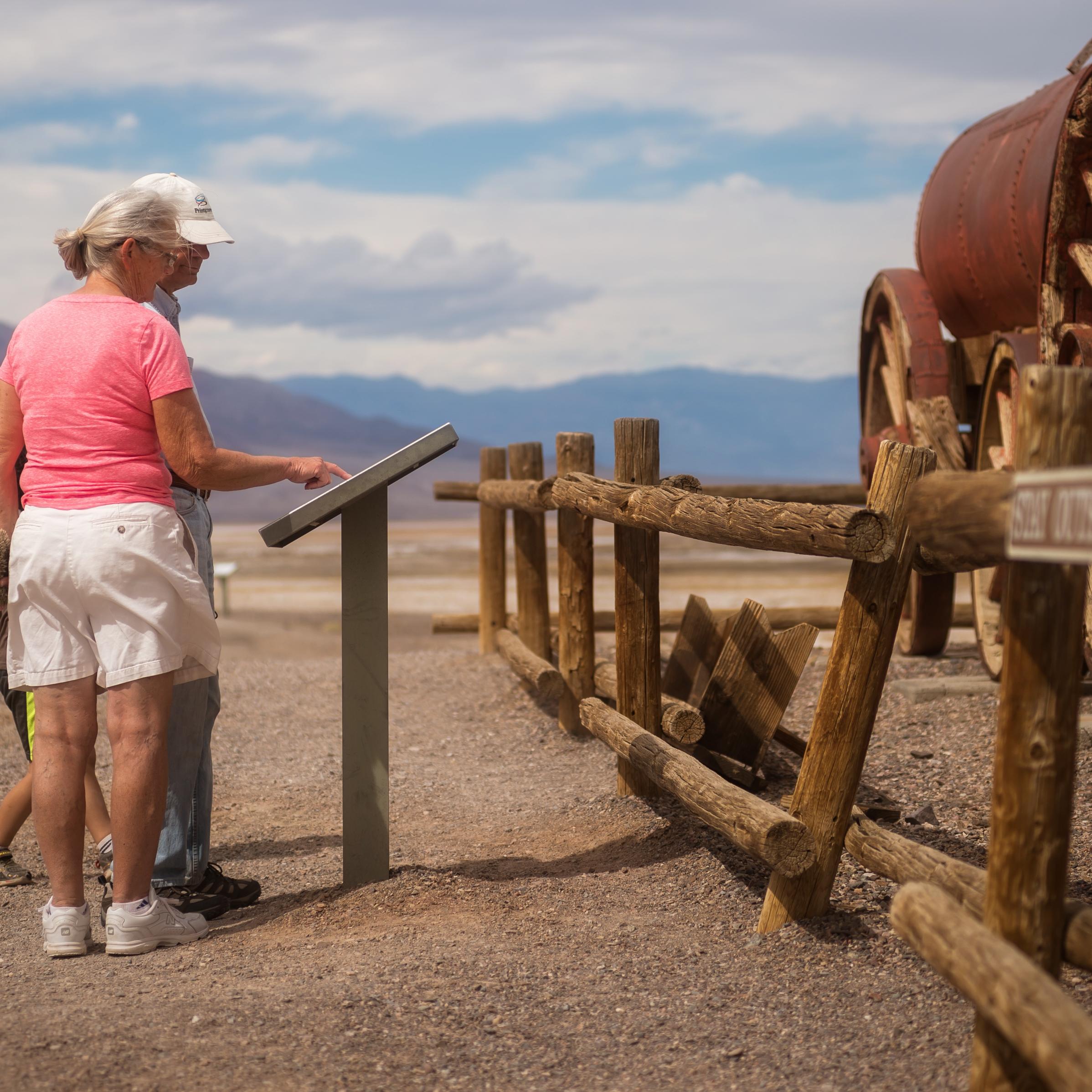 Two adults and a child look at a waist high sign next to a wooden and metal wagon in the desert.