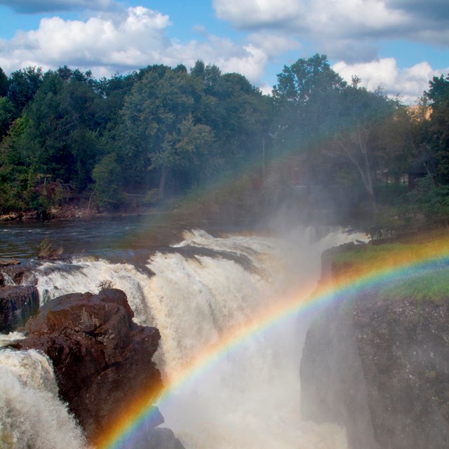 A rainbow over a grey rock cliff. Drops of water sparkle and splash as the water flows over the edge