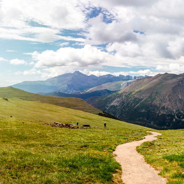 An alpine trail winds into the distance with panoramic view of craggy peaks.